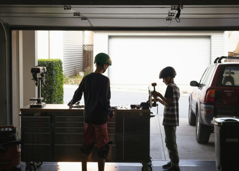 Two boys wearing helmets and standing in their garage with its door open, setting up a camera